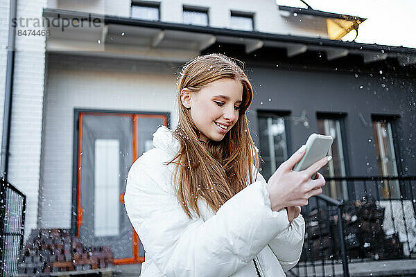 Glückliches Teenager-Mädchen  das außerhalb des Hauses Textnachrichten über das Smartphone sendet