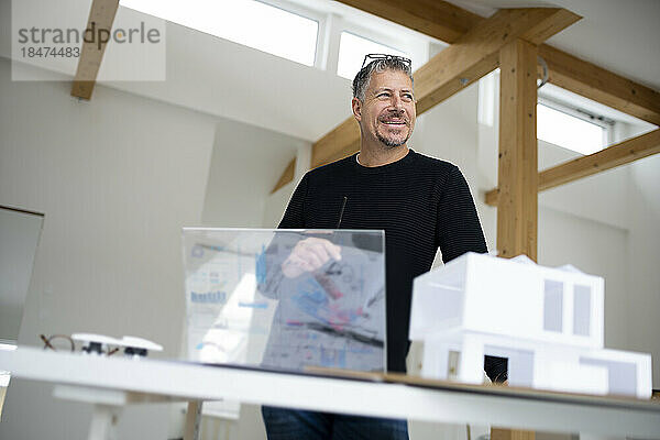 Lächelnder Architekt mit futuristischem Tablet-PC und Musterhaus am Schreibtisch