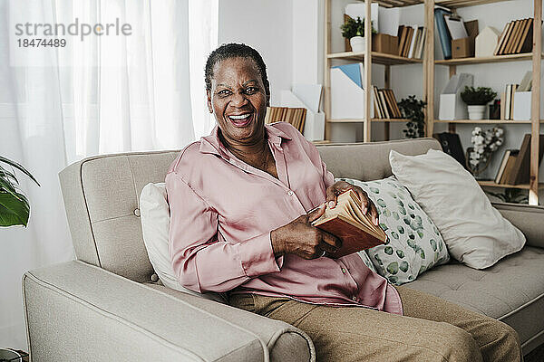 Glückliche ältere Frau mit Buch sitzt zu Hause auf dem Sofa