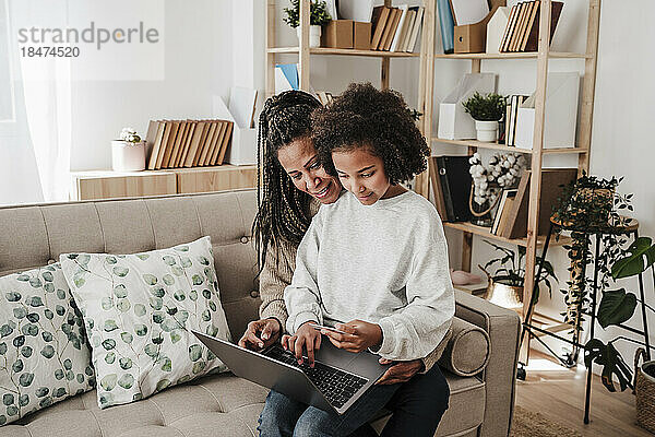 Tochter macht Online-Einkäufe mit Mutter über Laptop  die zu Hause auf dem Sofa sitzt