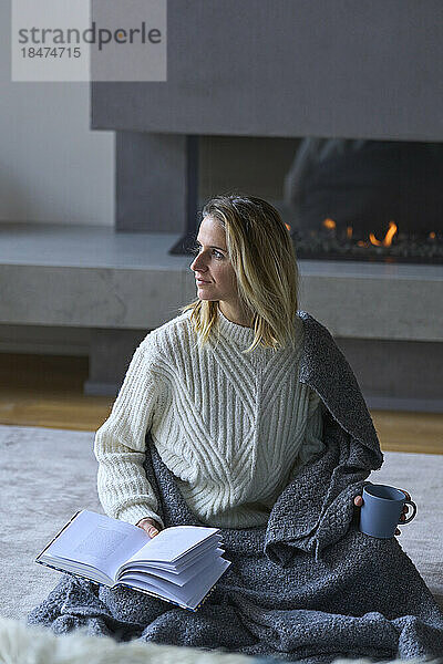 Nachdenkliche Frau  in eine Decke gehüllt  sitzt mit Buch und Kaffeetasse zu Hause