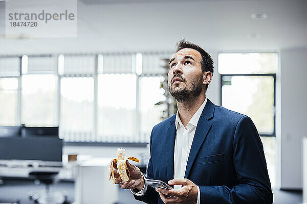 Geschäftsmann steht mit Banane und Smartphone im Büro