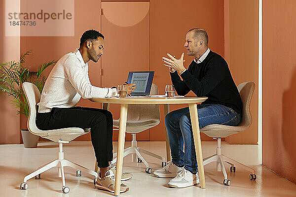 Geschäftsmann diskutiert mit Kollegen am Schreibtisch vor orangefarbener Wand