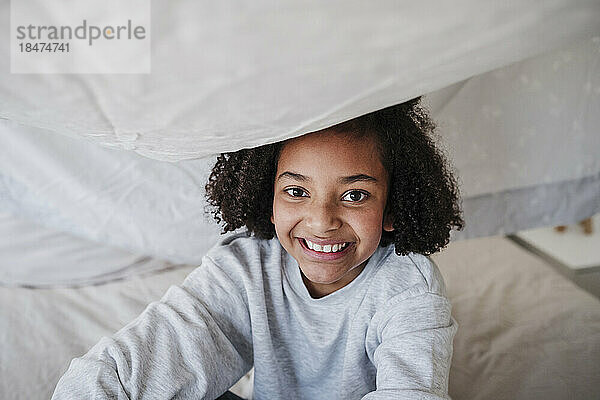 Glückliches Mädchen  das zu Hause unter einer Decke auf dem Bett sitzt