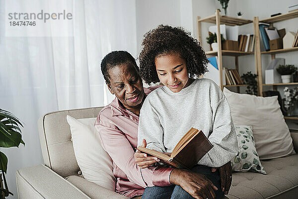 Enkelin liest Buch mit Großmutter  die zu Hause auf dem Sofa sitzt