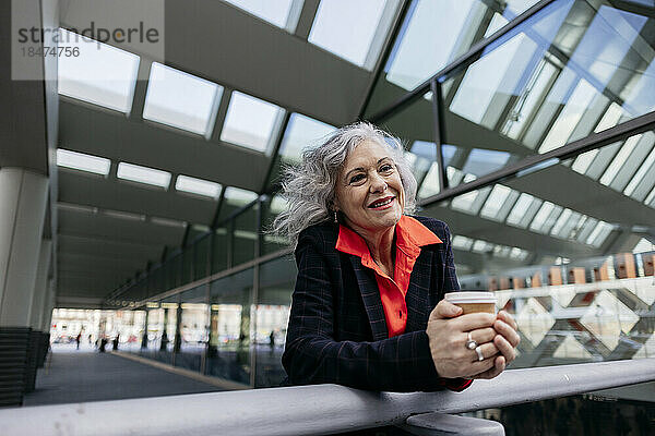 Lächelnde reife Geschäftsfrau mit Einwegkaffeetasse lehnt am Geländer