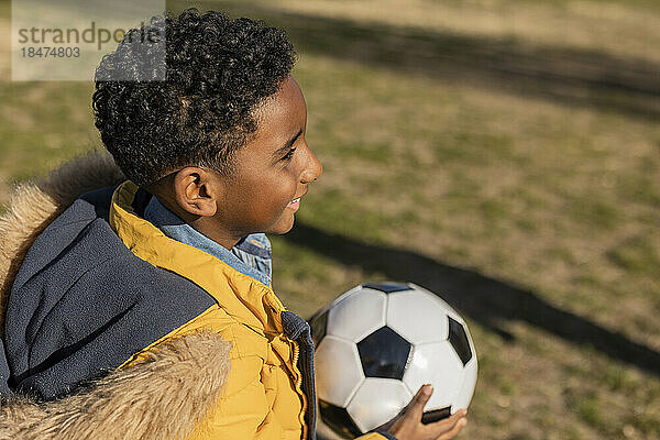 Fröhlicher Junge im Parka-Mantel hält Fußball im Park