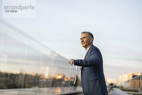 Smiling mature businessman standing by glass railing at sunset