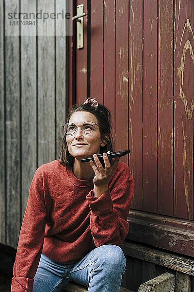 Lächelnde Frau mit Smartphone sitzt vor der Tür