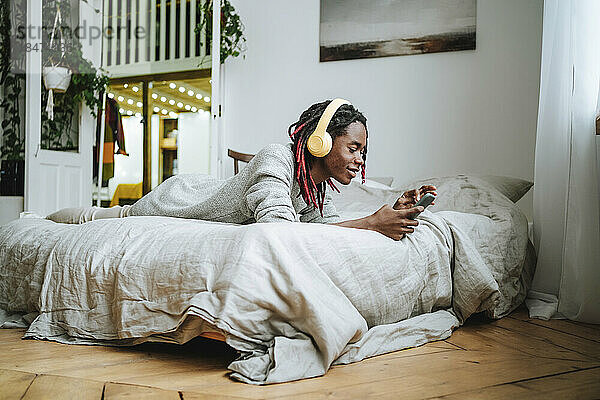 Lächelnder junger Mann mit Kopfhörern und Smartphone  der zu Hause im Bett liegt