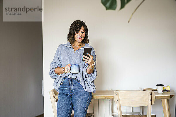Glückliche Frau mit Kaffeetasse und Smartphone zu Hause