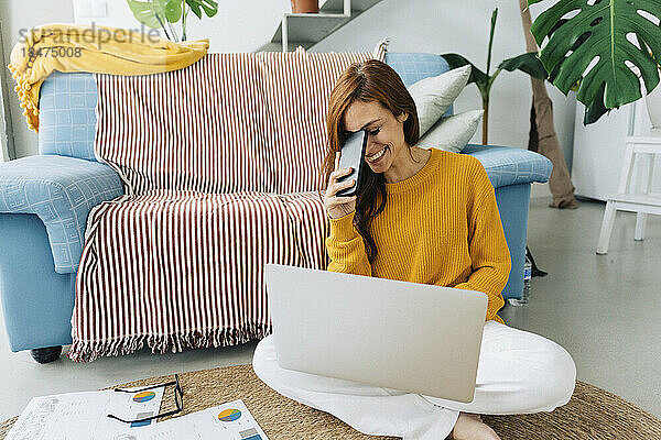 Glückliche Frau mit Smartphone und Laptop sitzt zu Hause auf dem Teppich