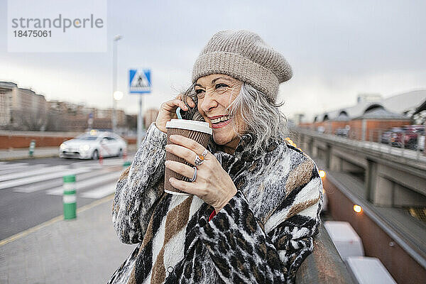 Glückliche reife Frau mit Einwegbecher  die auf dem Fußweg mit dem Smartphone spricht