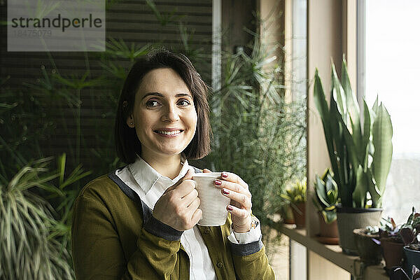 Lächelnde Frau hält Kaffeetasse vor Pflanzen