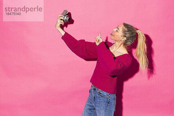 Frau zeigt Frieden und macht vor rosa Hintergrund ein Selfie mit der Kamera
