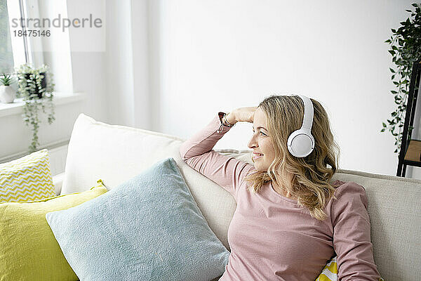 Nachdenkliche reife Frau mit kabellosen Kopfhörern sitzt zu Hause