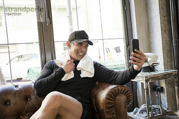 Glücklicher Mann  der auf dem Sofa im Fitnessstudio sitzt und ein Selfie mit dem Smartphone macht