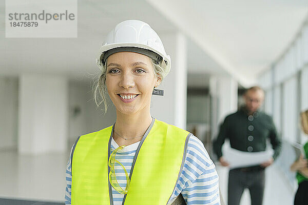 Lächelnde Frau in Schutzkleidung mit Kollegen im Hintergrund im Büro