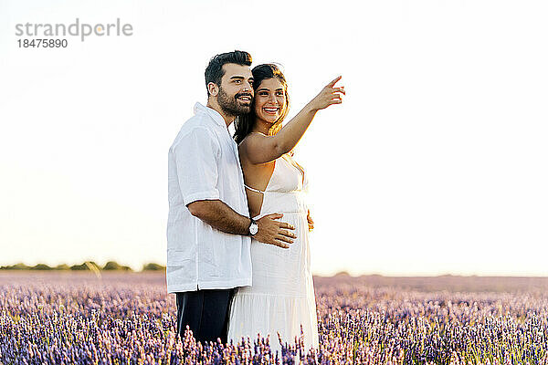 Glückliche Frau zeigt in die Ferne und steht mit Mann im Lavendelfeld bei Sonnenuntergang