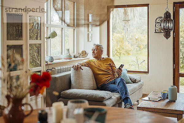 Nachdenklicher älterer Mann sitzt mit Smartphone zu Hause auf dem Sofa