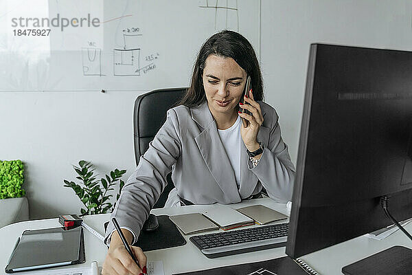 Lächelnde Geschäftsfrau  die am Schreibtisch im Büro sitzt und mit einem Smartphone spricht