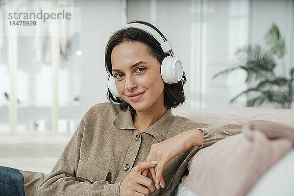 Lächelnde Frau mit kabellosen Kopfhörern sitzt im Schlafzimmer