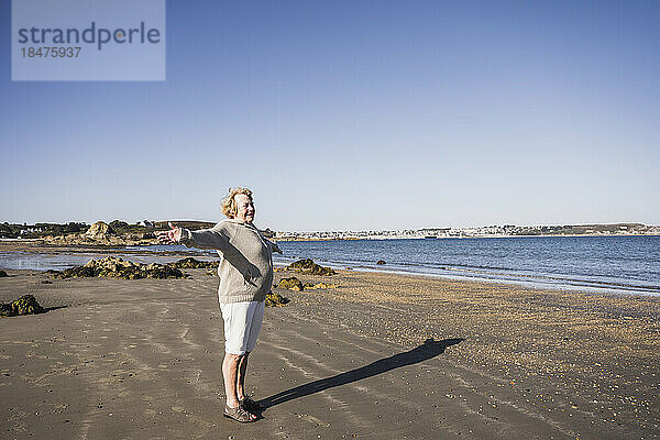 Sorglose ältere Frau mit ausgestreckten Armen steht am Strand
