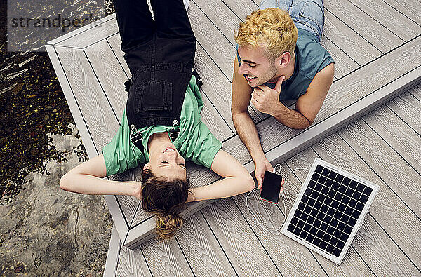 Lächelndes Paar entspannt auf der Terrasse mit Smartphone  das über ein Solarpanel aufgeladen wird