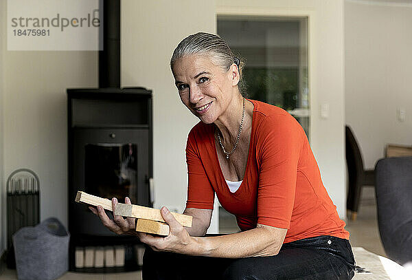 Lächelnde Frau mit Brennholz sitzt zu Hause im Wohnzimmer