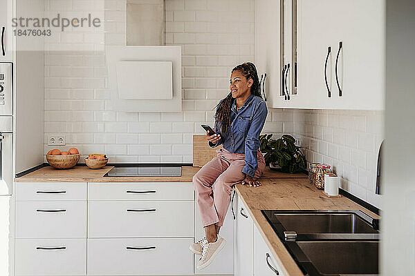 Lächelnde Frau sitzt zu Hause mit Smartphone auf der Küchentheke