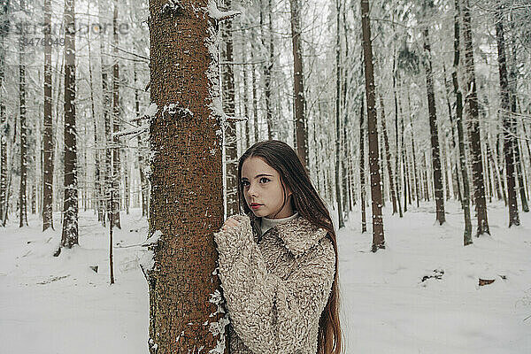 Nachdenkliches Teenager-Mädchen steht am Baum im verschneiten Wald