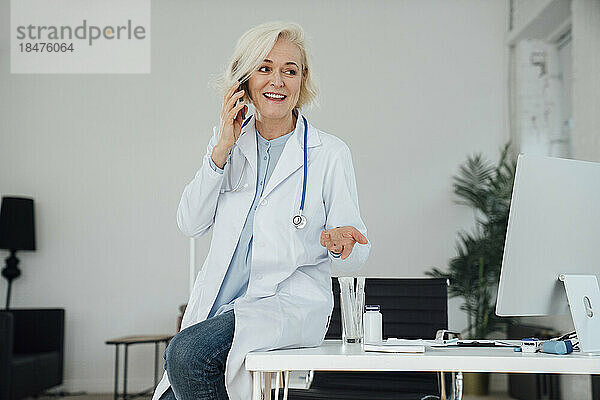 Lächelnder reifer Arzt  der in der Klinik auf dem Smartphone spricht