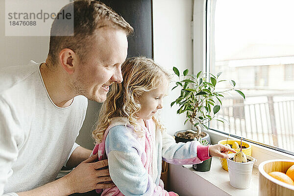 Happy father and daughter taking care of plants on window sill at home