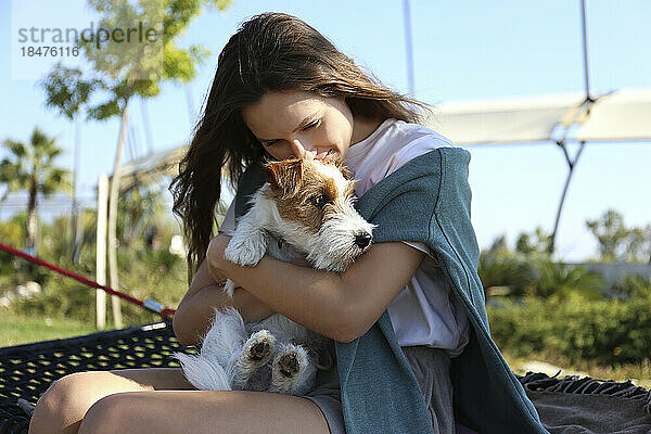 Glückliche junge Frau mit Jack Russell Terrier sitzt an einem sonnigen Tag in der Hängematte