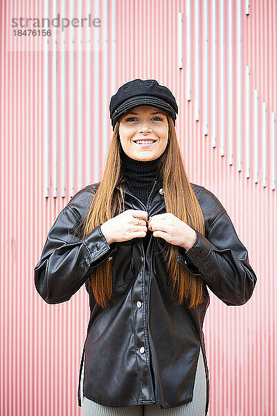 Glückliche rothaarige Frau mit Mütze und Jacke vor rosa Wand