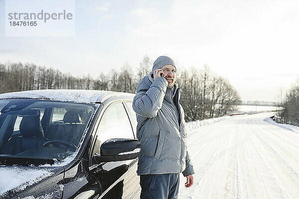 Lächelnder Mann in warmer Kleidung  der vor dem Auto auf der winterlichen Straße mit dem Smartphone spricht