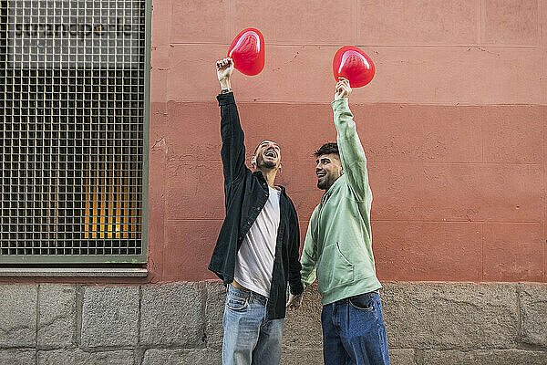 Glücklicher schwuler Mann mit Freund  der einen roten Ballon vor der Wand hält