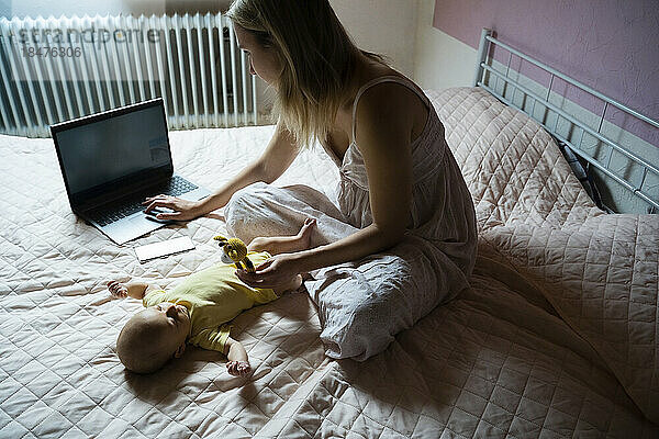 Berufstätige Mutter benutzt Laptop und spielt mit kleinem Jungen  der zu Hause im Bett liegt