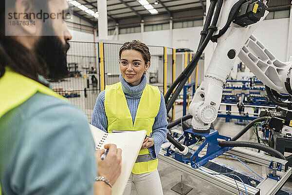 Lächelnder Ingenieur blickt auf Kollegen mit Notizblock  der neben Roboterarm steht