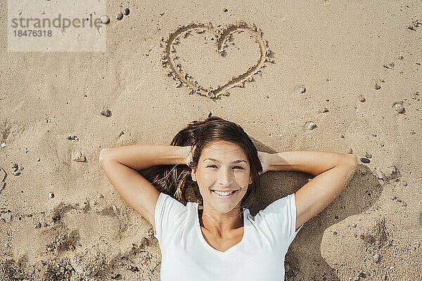 Lächelnde Frau mit den Händen hinter dem Kopf  die am Strand im Sand liegt