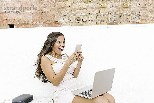 Überraschte Frau benutzt Smartphone und sitzt mit Laptop auf Bank