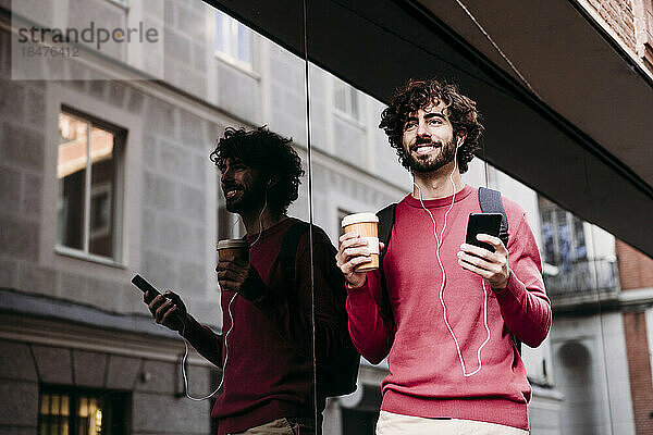Lächelnder junger Mann mit Smartphone und Kaffeetasse steht am Fußweg
