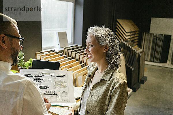 Lächelnde Geschäftsfrau blickt auf Kollegen  der im Architekturbüro einen Bauplan hält