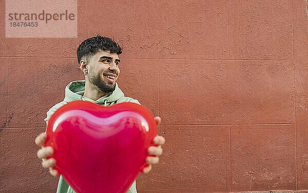 Happy young man holding red heart shape balloon in front of wall