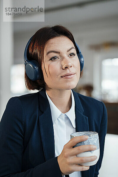 Nachdenkliche Geschäftsfrau hört im Büro Musik über Kopfhörer mit Kaffeetasse
