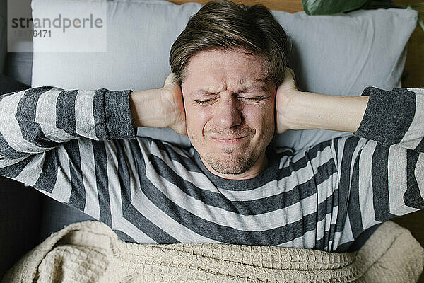 Frustrated man covering ears lying on bed at home