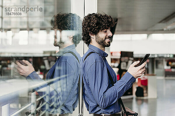 Lächelnder Geschäftsmann lehnt an Glas und benutzt Smartphone am Bahnhof