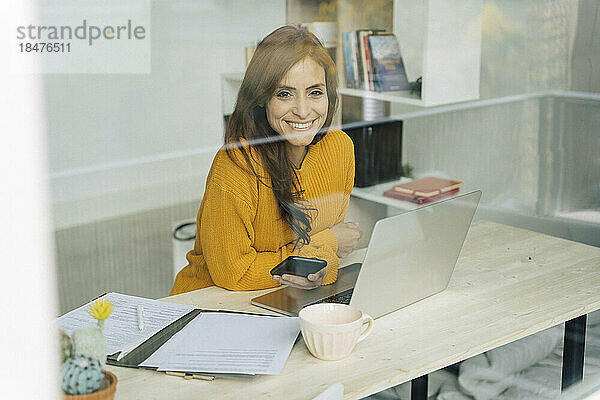 Glückliche Freiberuflerin mit Smartphone und Laptop am Schreibtisch