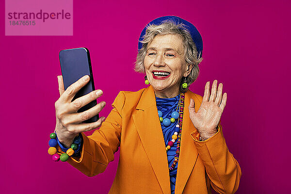 Glückliche ältere Frau winkt bei einem Videoanruf über ihr Smartphone vor rosa Hintergrund