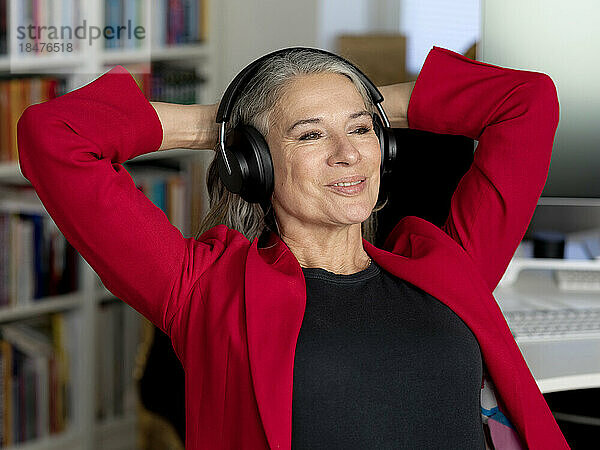 Glückliche Geschäftsfrau mit kabellosen Kopfhörern sitzt mit den Händen hinter dem Kopf